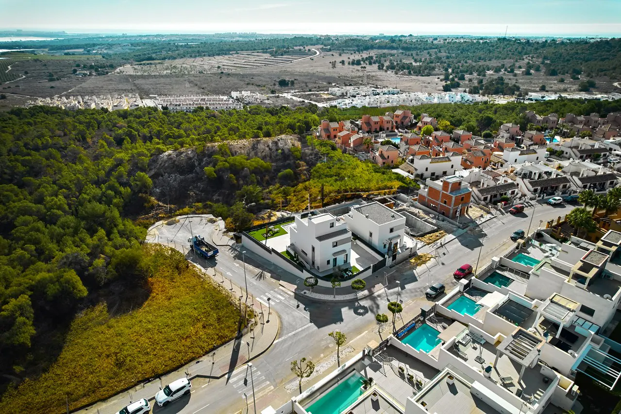 Aerial view of a modern residential area with white houses, pools, green trees, and distant landscape.