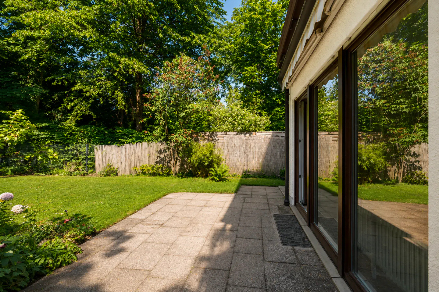 A paved patio leads to a lush green backyard with a wooden fence, surrounded by mature trees and flowering plants.