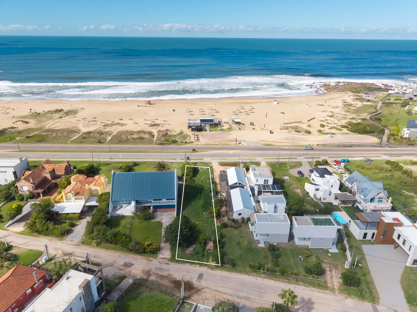 Aerial view of a vacant lot outlined in white, near a sandy beach and blue ocean. Houses line the street.