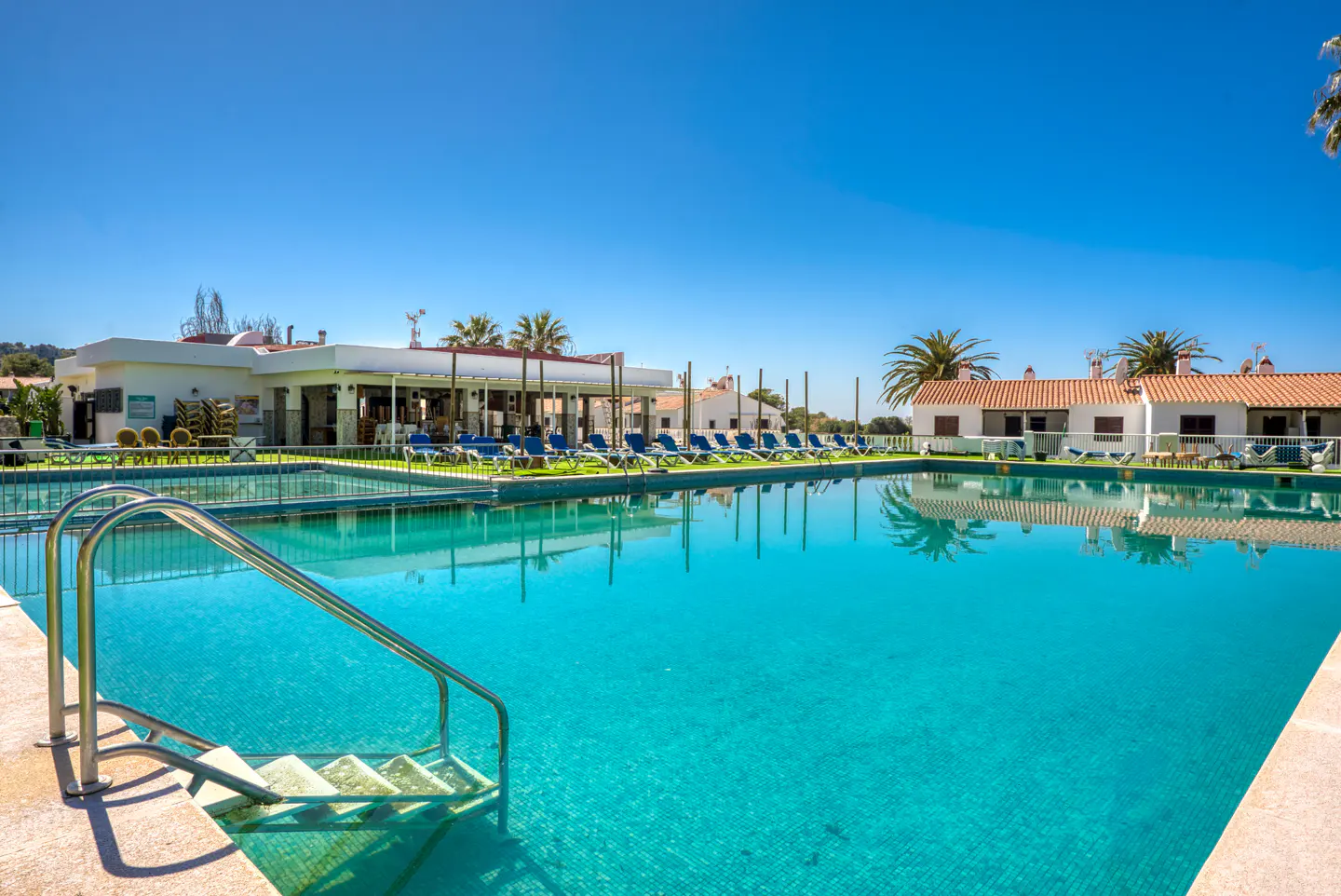 A bright blue swimming pool with metal steps, surrounded by lounge chairs and white buildings under a clear blue sky.