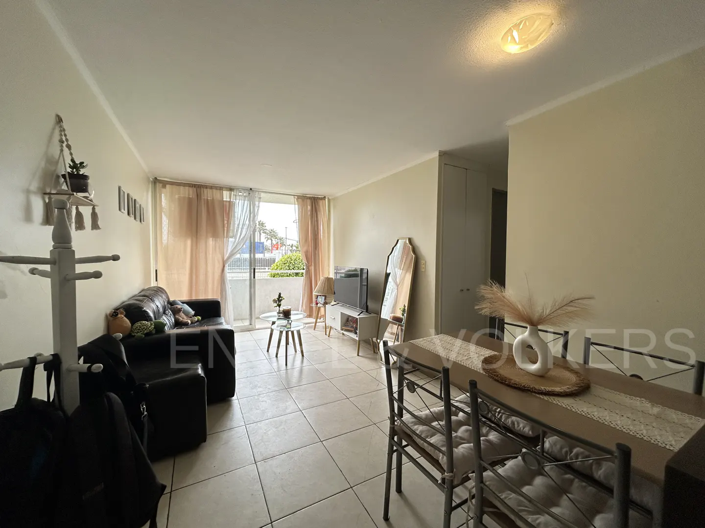 Bright living room with tile floors, black leather sofa, dining table with chairs, and a balcony with sheer curtains.