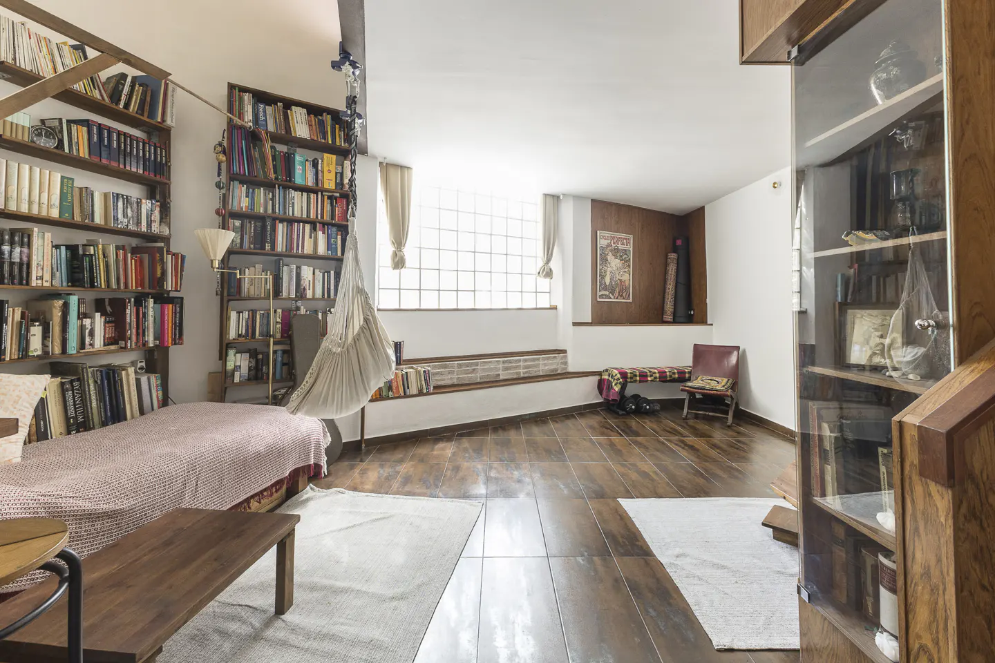 Cozy living room with floor-to-ceiling bookshelves, a hammock, and a window seat. Hardwood floors and a glass-fronted cabinet add warmth.