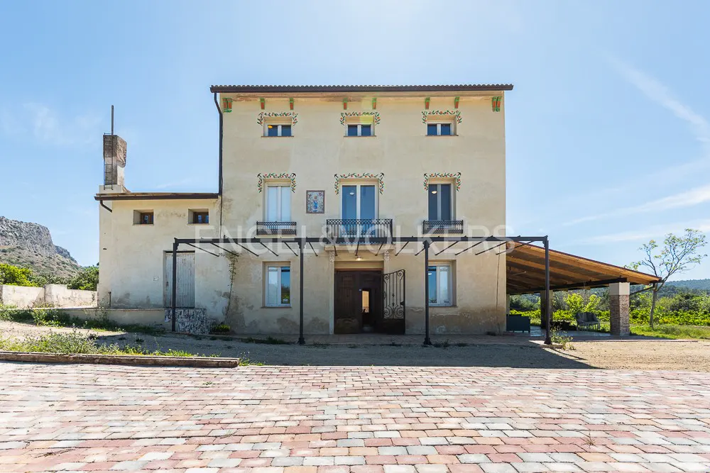 Two-story beige house with a balcony, decorative trim, and a brick driveway under a clear blue sky.