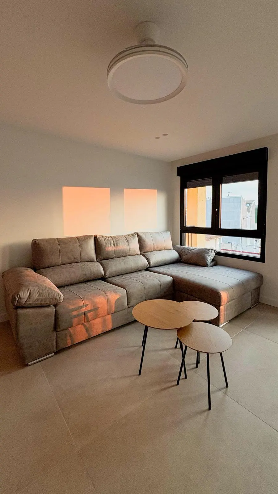 Living room with a large gray sectional sofa, two wooden coffee tables, and a window with black frames. Sunlight shines on the wall.