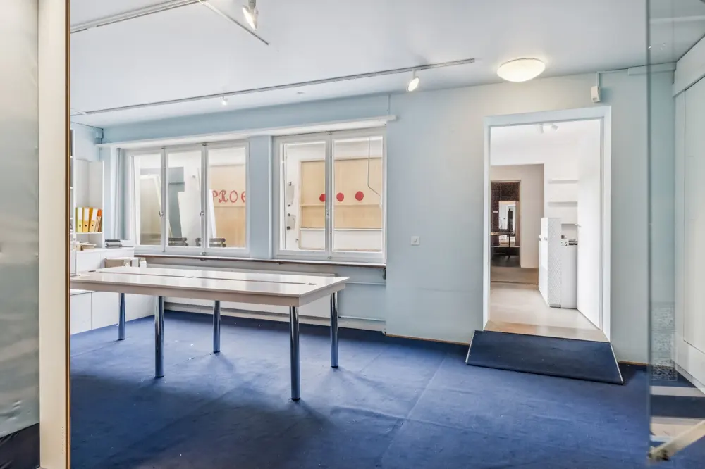 Empty office space with blue carpet, white walls, and a table. Windows let in natural light. A doorway leads to another room.
