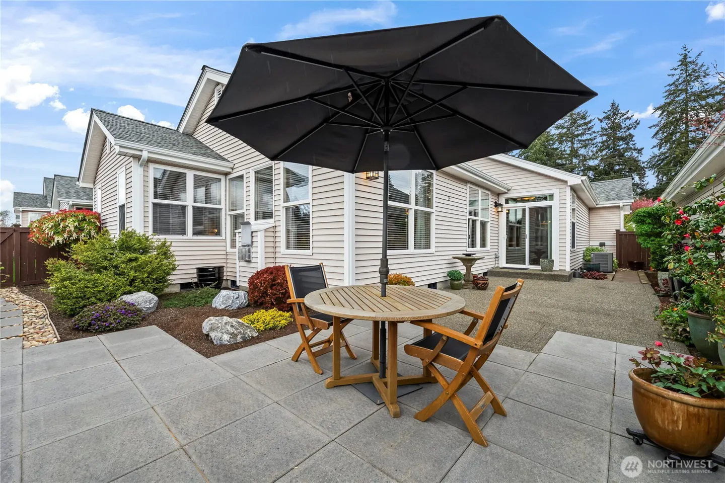 Backyard patio with a round wooden table, chairs, and a black umbrella. Landscaping and siding of the house are visible.