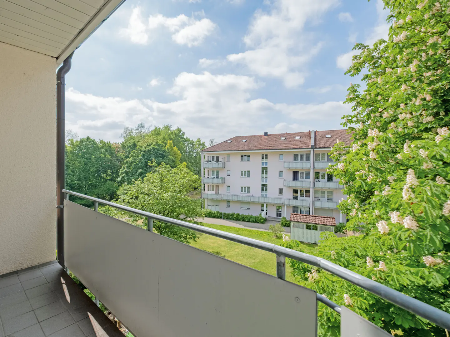 View from a balcony overlooking a green lawn, lush trees, and a multi-story apartment building under a blue sky.
