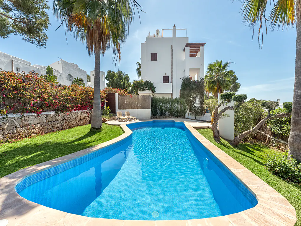 A rectangular blue pool is surrounded by green grass, palm trees, and a white building under a clear blue sky.