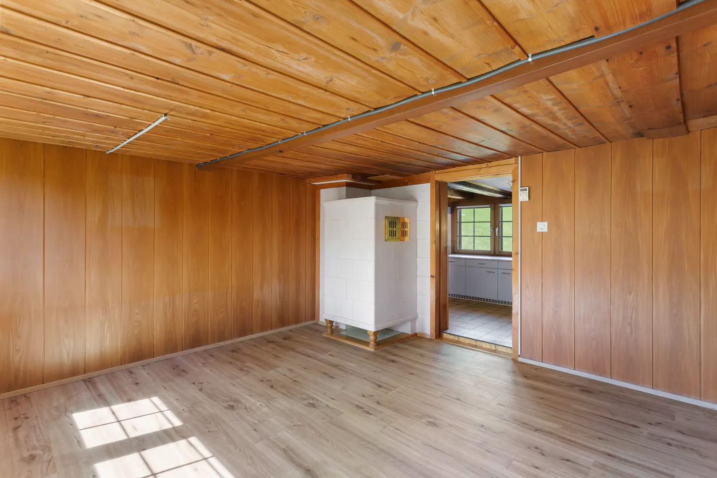 An empty room with wood-paneled walls, ceiling, and light wood floors. A white stove is in the corner, and a doorway leads to a kitchen.