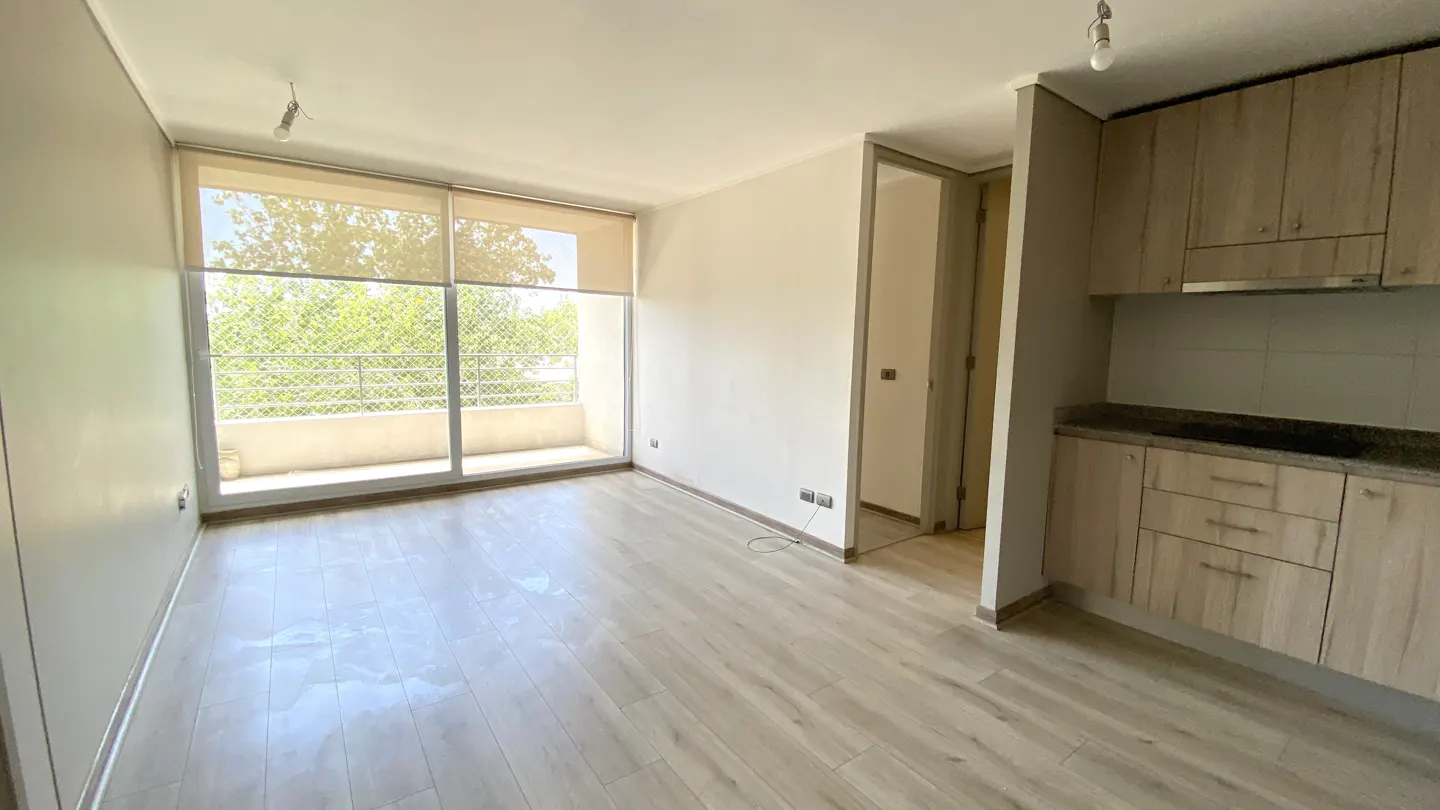 Bright, empty apartment with wood floors, a balcony with a view, and a kitchen with light wood cabinets.
