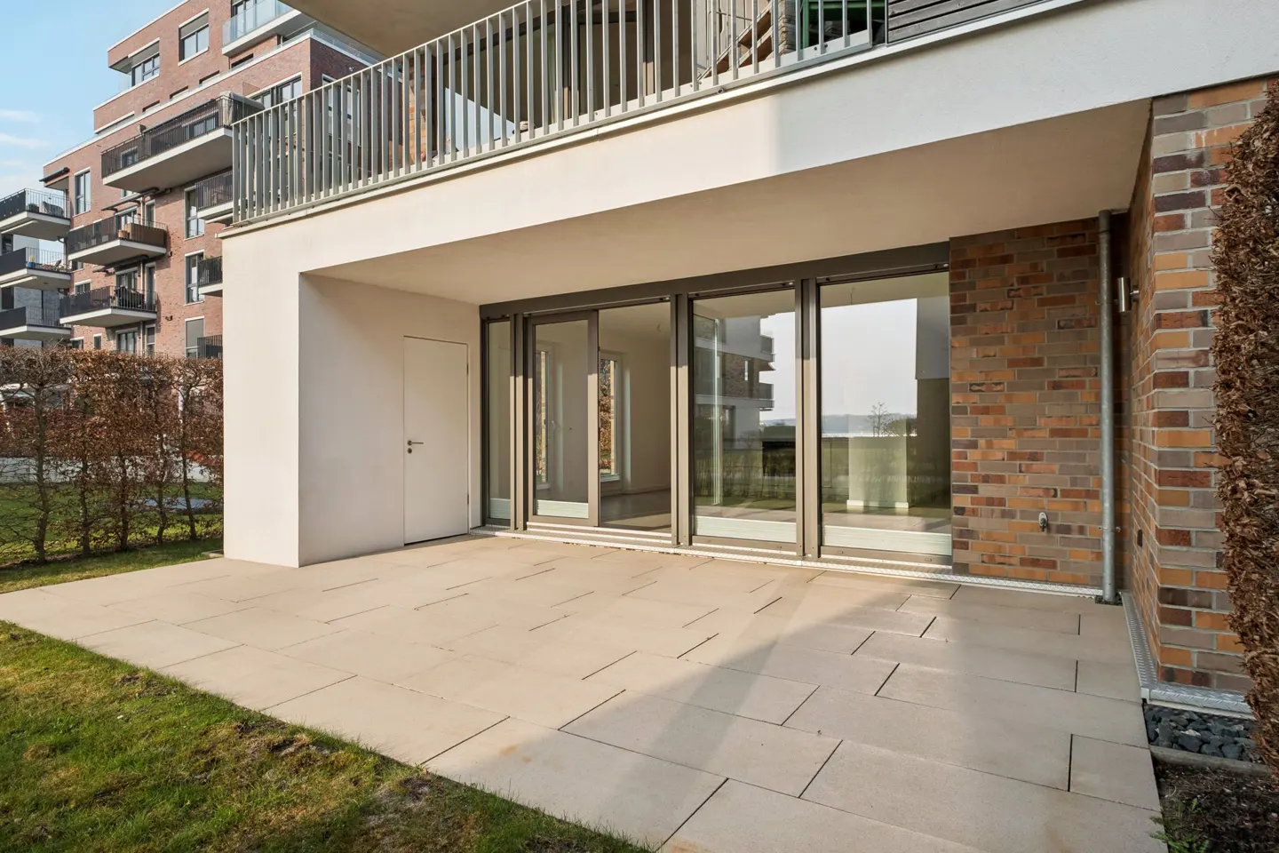 Exterior view of a modern apartment patio with sliding glass doors, a white door, and a stone-paved patio. A brick wall and green grass surround the patio.