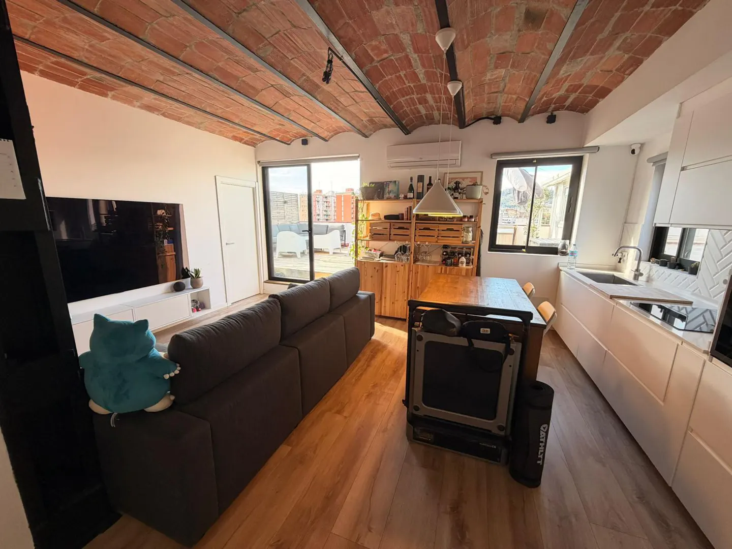 Bright, modern apartment with brick ceiling, wood floors, and gray sofa. White kitchen cabinets and a dining table are visible.