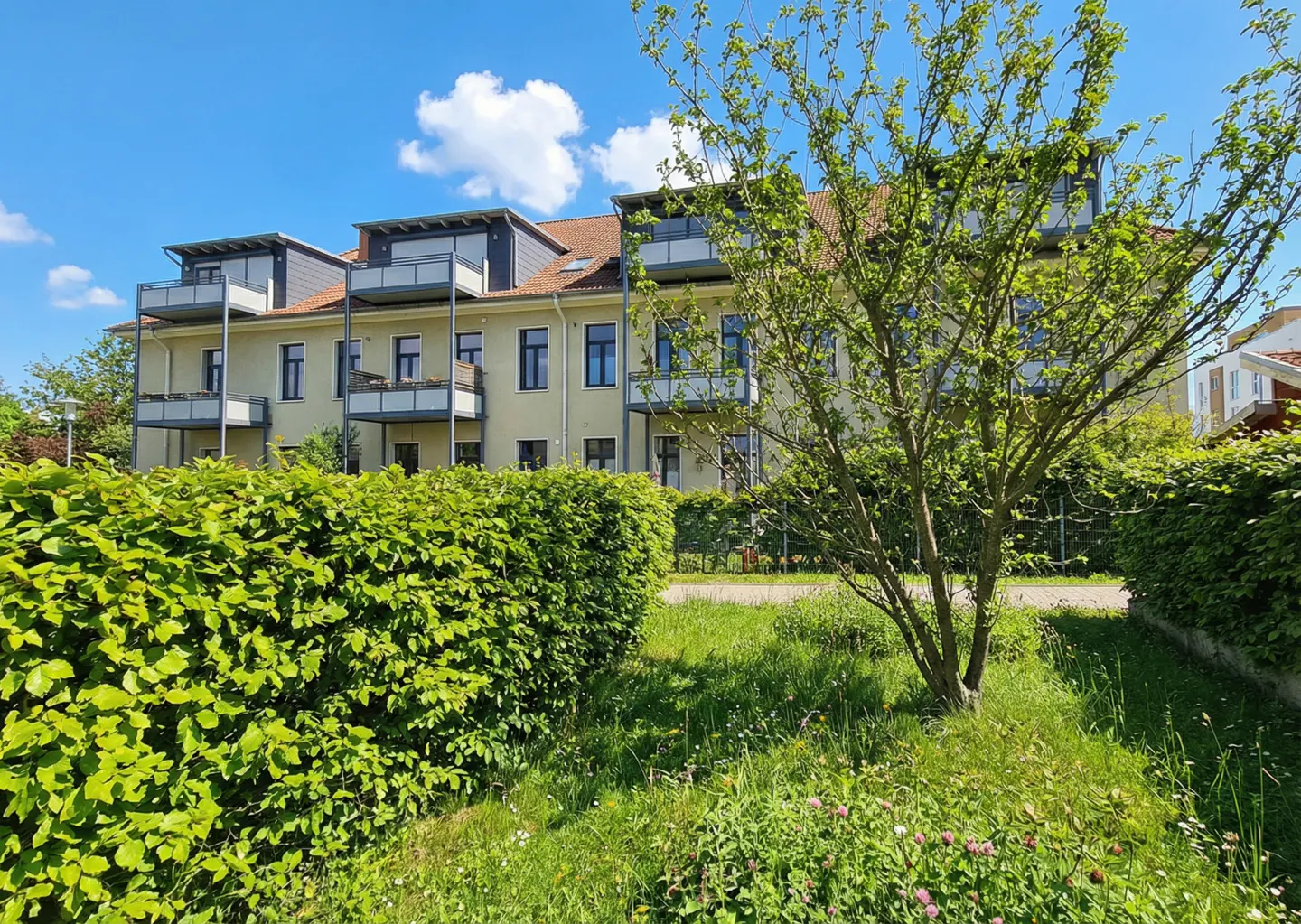 Exterior view of a three-story apartment building with balconies, green hedges, and a tree in the foreground under a blue sky.