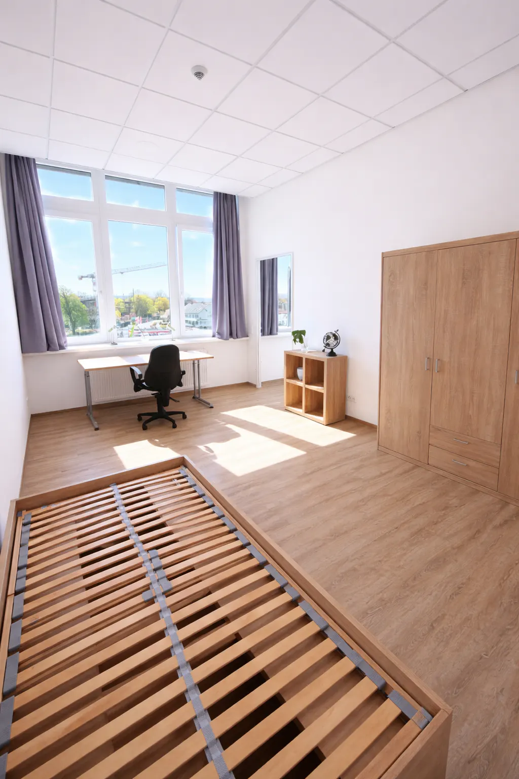 Bright, empty bedroom with a wooden bed frame, desk, chair, and wardrobe. Large windows with gray curtains let in natural light.