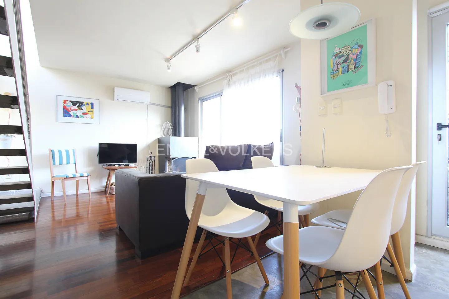 Bright living room with hardwood floors, white walls, and a modern white table and chairs. A gray sofa and TV are in the background.