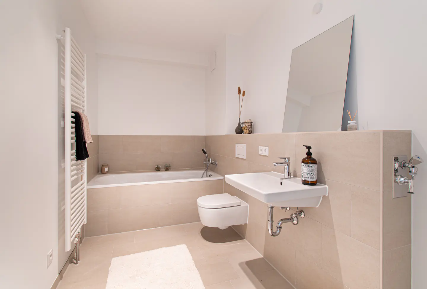 Bright bathroom with beige tile, white walls, and modern fixtures. A white sink, toilet, and bathtub are visible. A towel warmer stands to the left.