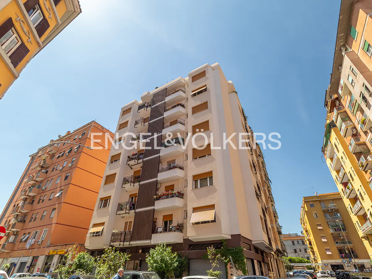 Exterior view of a tall, multi-story apartment building with balconies, surrounded by other buildings under a clear blue sky.