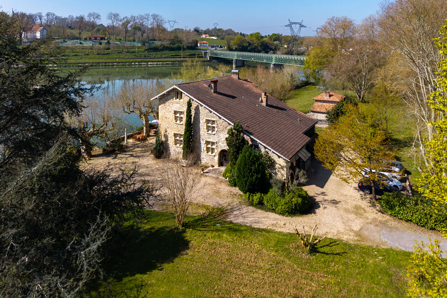 Aerial view of a stone house with a brown roof, near a river and a green bridge, surrounded by trees and a green lawn.