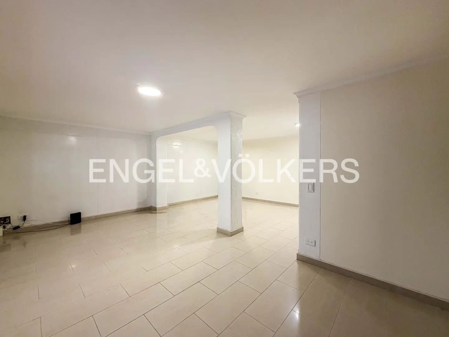 An empty room with white walls, beige tile floors, and white pillars. Engel & Völkers logo is superimposed on the image.