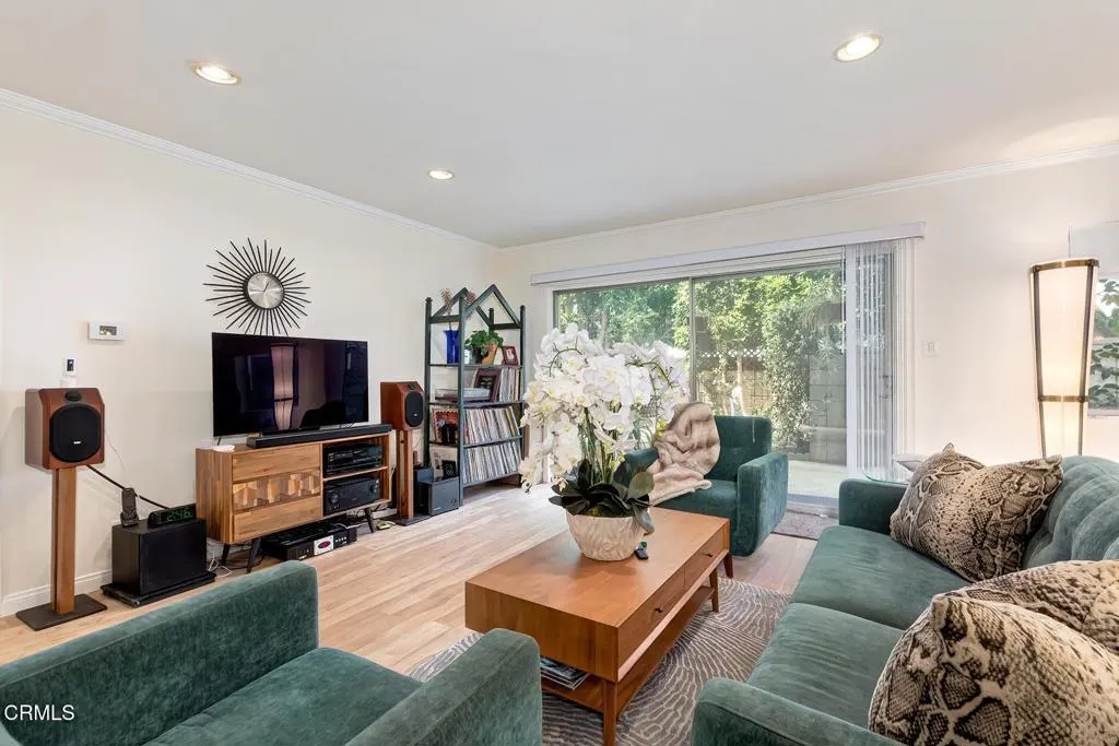 Living room with green sofas, wood floors, and a sliding glass door to a green backyard. A TV and speakers are on a wooden console.