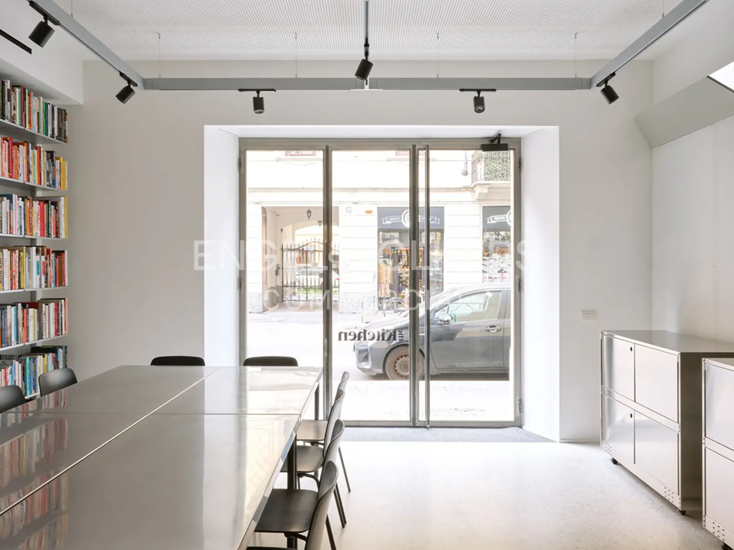 Bright, modern office space with a long table, chairs, and a bookshelf. A glass door shows a street view with a car.