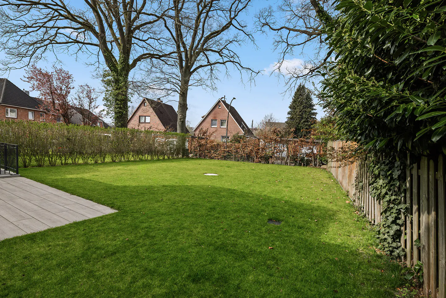 A lush green lawn with a wooden fence and trees in the background. Brick houses are visible beyond the hedge.