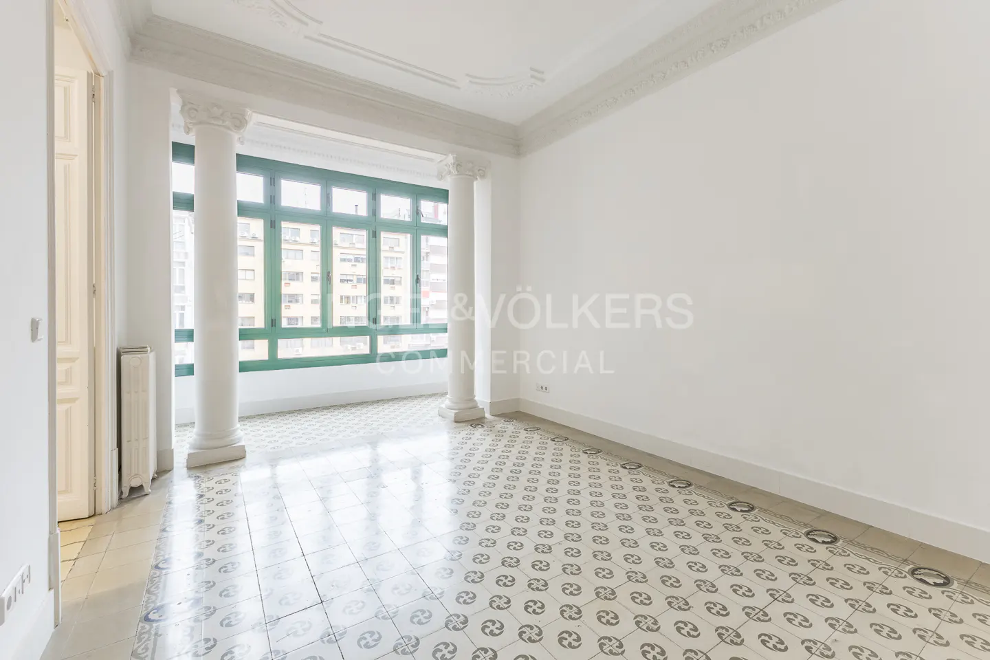 Bright, empty room with white walls, patterned tile floor, and green-framed windows with white columns.