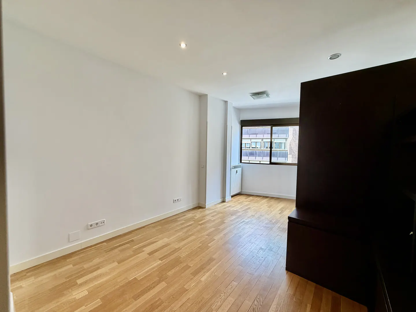 Bright, empty room with light wood floors, white walls, and a window with black frame. A dark brown cabinet is on the right.
