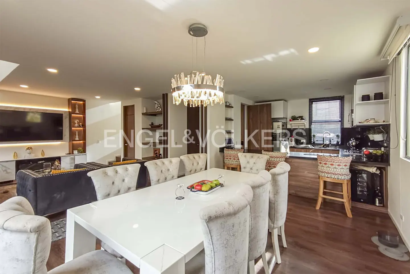 Open-concept living and dining area with a white table, upholstered chairs, and a modern chandelier. Kitchen and living room in the background.