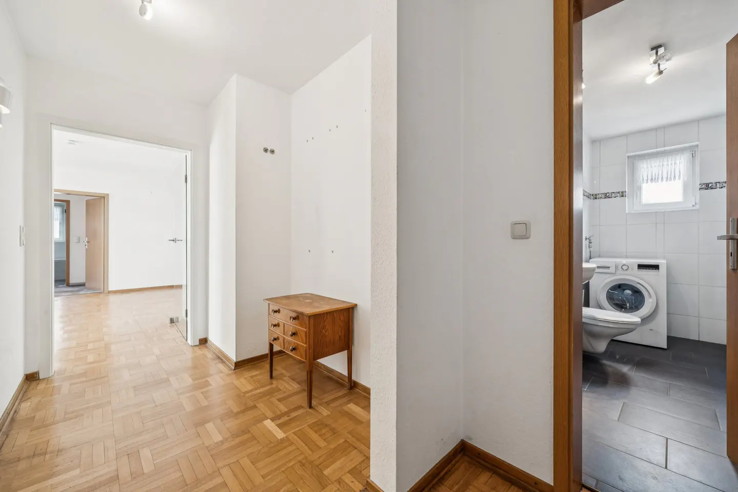 Hallway with parquet flooring, a small wooden chest of drawers, and a view into a tiled bathroom with a washing machine.