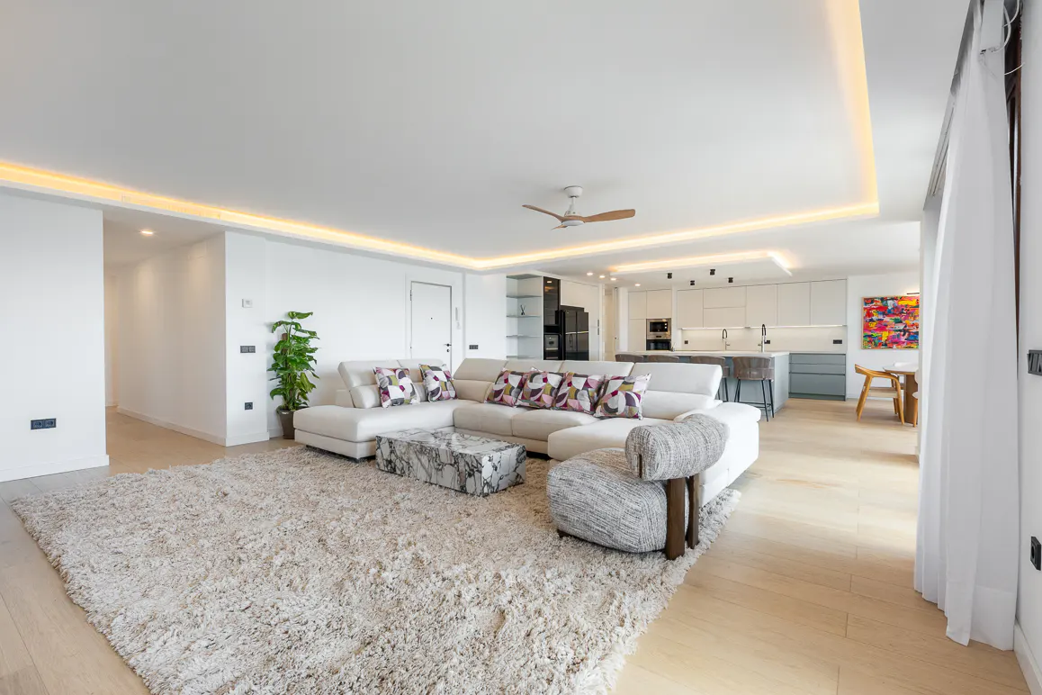 Bright, open-concept living space with a white sectional sofa, marble coffee table, and a plush rug. Kitchen in the background.
