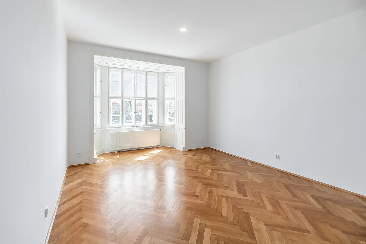 Bright, empty room with herringbone wood floor, white walls, and a large bay window with a radiator.