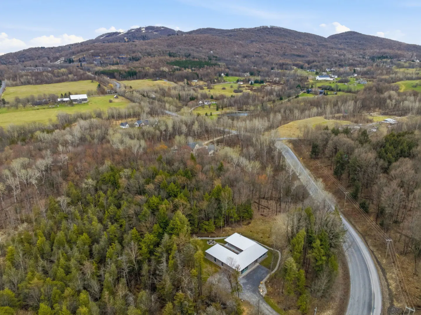 Aerial view of a modern, U-shaped house with a gray roof, surrounded by trees, a winding road, and distant mountains under a blue sky.