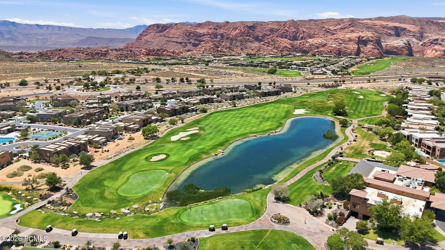 Aerial view of a lush green golf course with a large blue pond, surrounded by desert homes and red rock mountains.