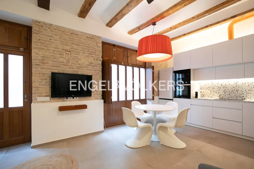 A modern apartment interior with exposed brick, wooden beams, and a white kitchen. A round table with white chairs sits under a red pendant light.