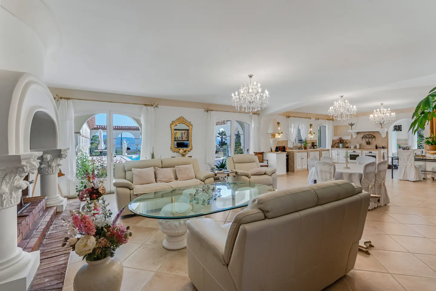 Bright, open-concept living room with beige sofas, glass coffee table, and chandeliers. Arched windows offer views of a pool and the sea.