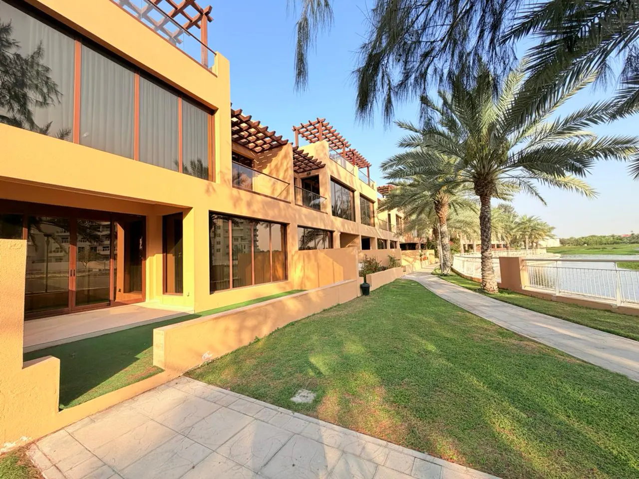 Exterior view of tan townhouses with large windows, balconies, and palm trees along a grassy walkway near a lake.
