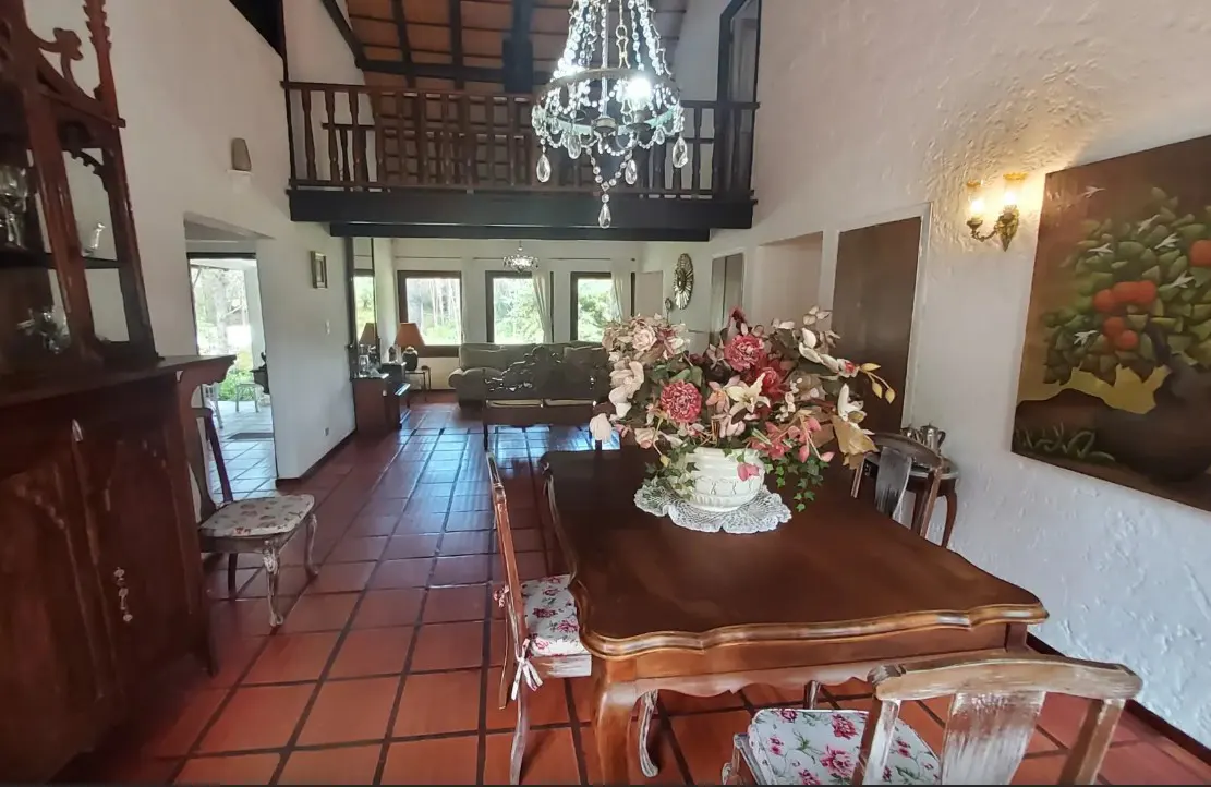A dining room with a wooden table, chairs, and a floral centerpiece. A chandelier hangs above, and a balcony overlooks the space.