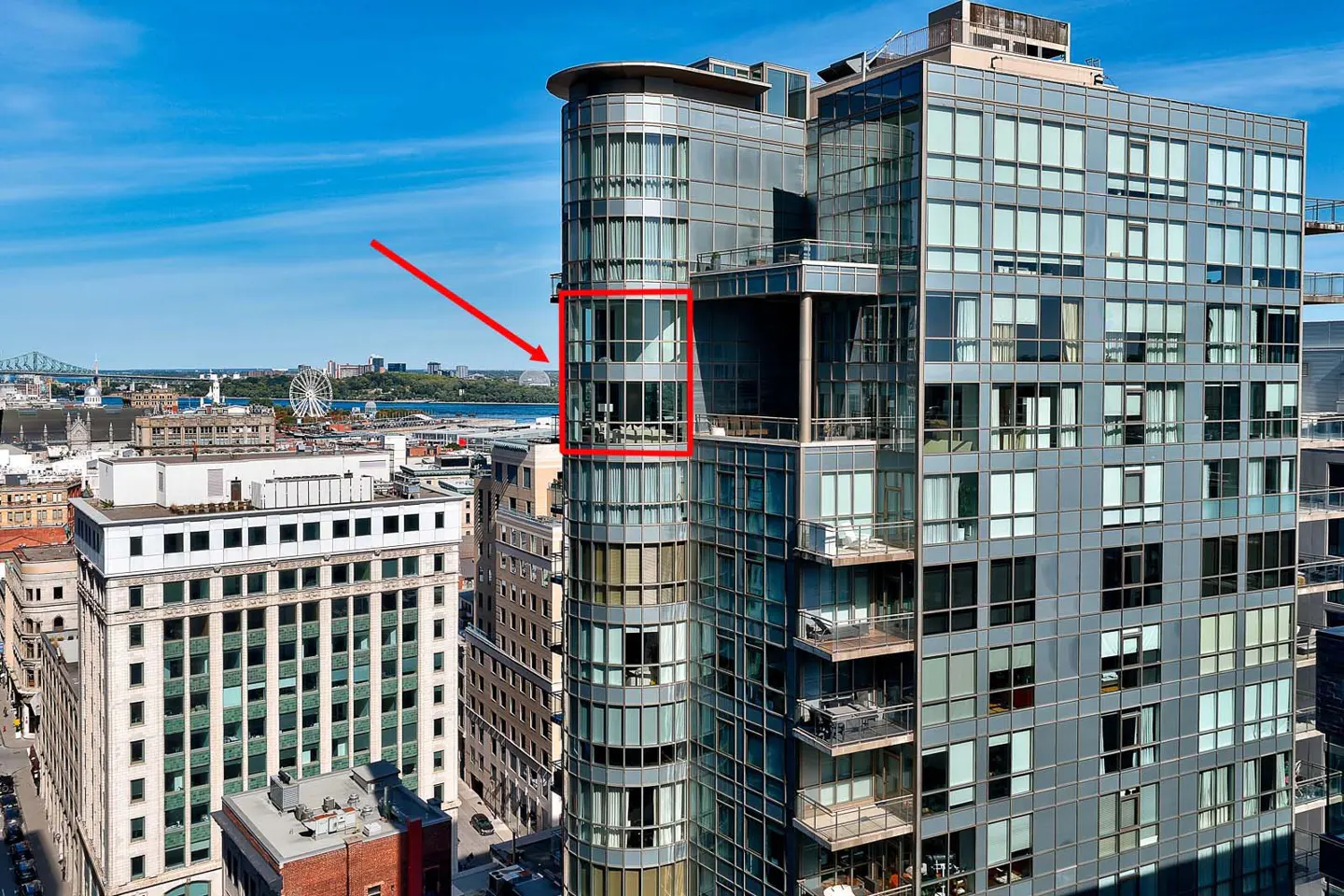 Exterior view of a modern high-rise condo with a balcony, indicated by a red arrow, overlooking a cityscape with a river and bridge.