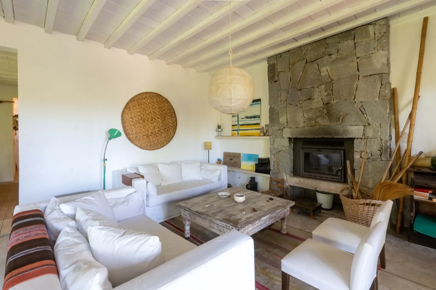 Living room with white sofas, stone fireplace, and rustic wood table on a striped rug. A woven basket hangs on the wall.