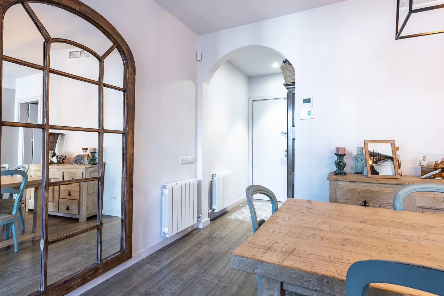 A bright dining room with a wooden table, blue chairs, and a large arched mirror reflecting the room. An arched doorway leads to a hallway.