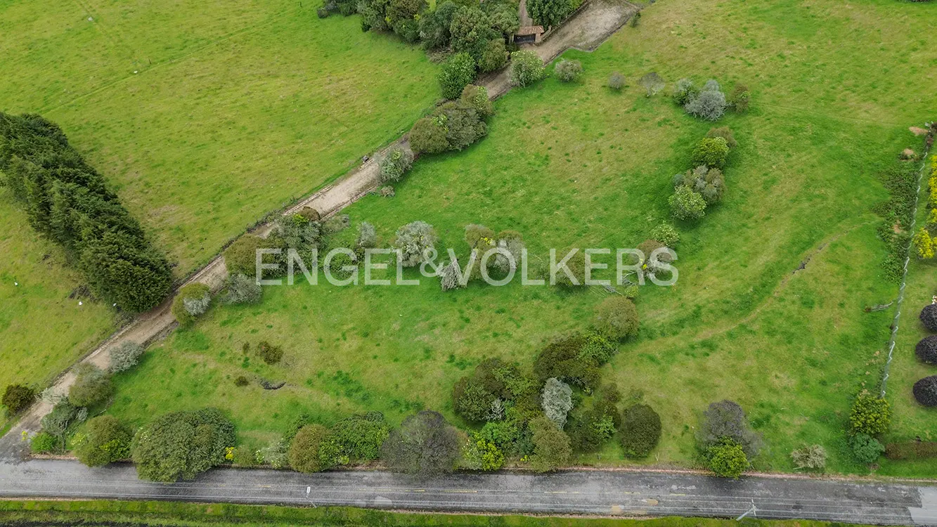 Aerial view of a green field with trees, a dirt path, and a road. The Engel & Volkers logo is superimposed on the image.