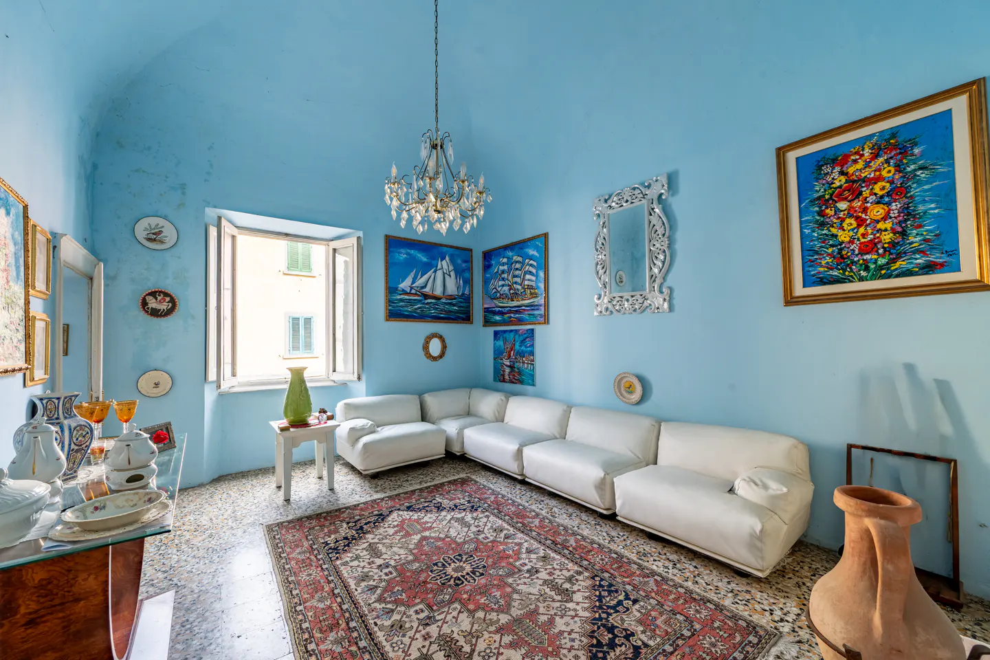 A light blue living room with a white sectional sofa, a patterned rug, and paintings on the walls. A chandelier hangs from the ceiling.