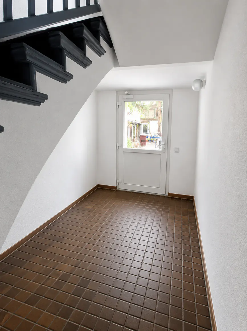 Entryway with brown tiled floor, white walls, and a white door with a window. A black staircase is visible on the left.