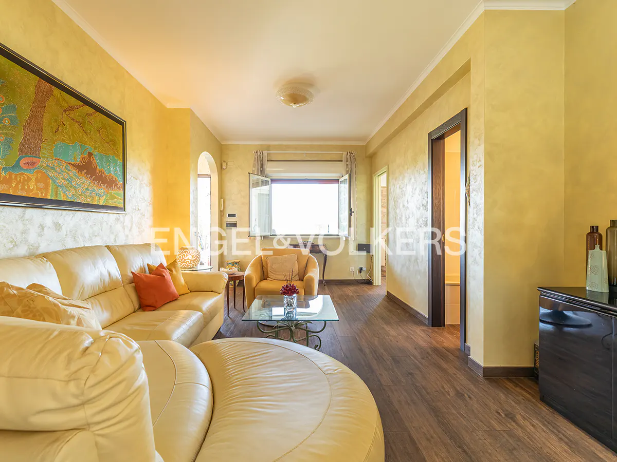Bright living room with yellow walls, wood floors, and a large window. A cream-colored sofa and armchair are arranged around a glass coffee table.
