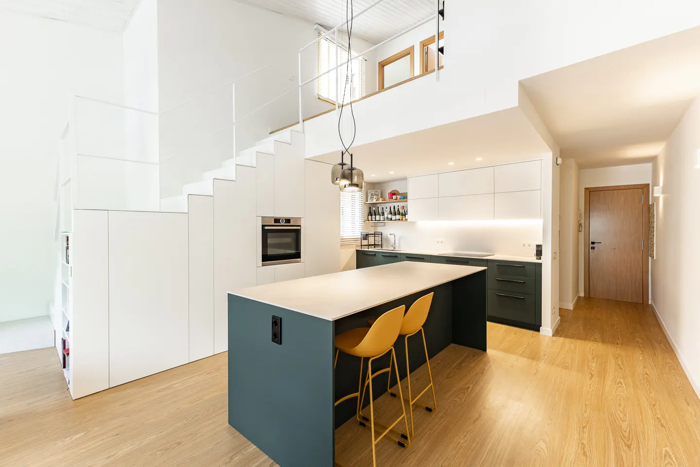 Modern kitchen with a dark teal island, two yellow bar stools, white cabinets, and a staircase leading to a loft area.