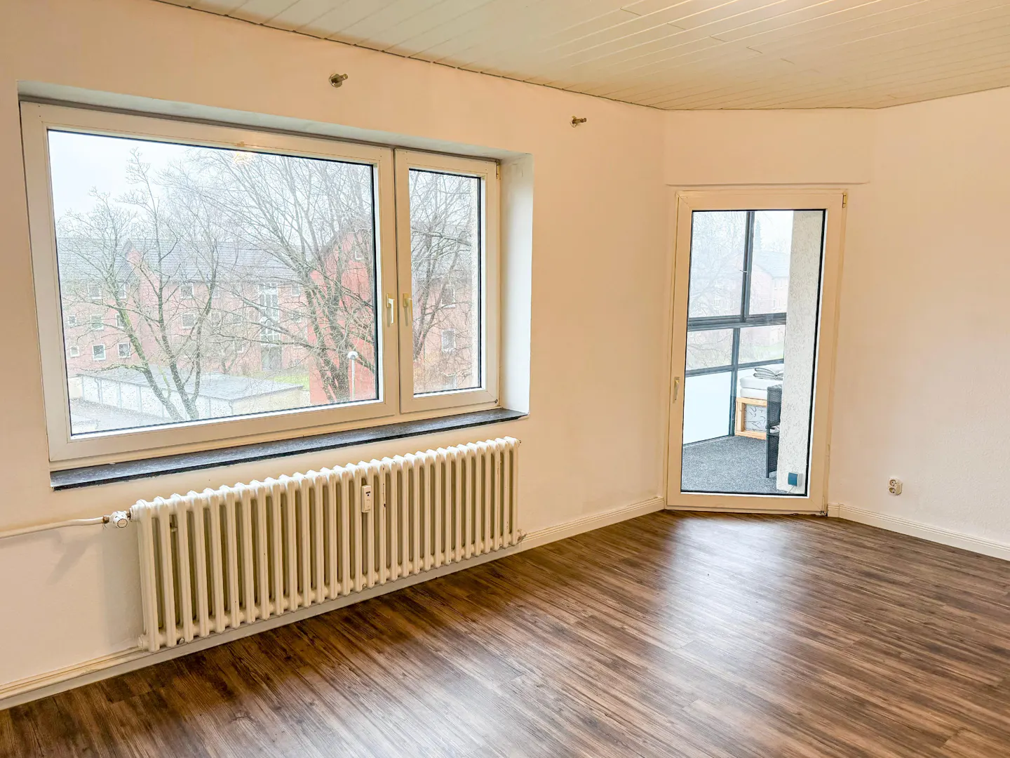 Bright, empty room with wood floors, white walls, and a radiator under a large window. A glass door leads to a balcony.