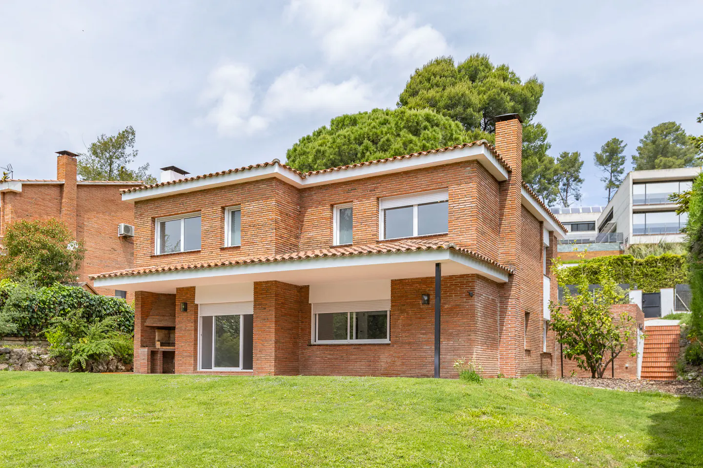 Two-story brick house with a red tile roof and white trim, set on a green lawn with trees in the background.