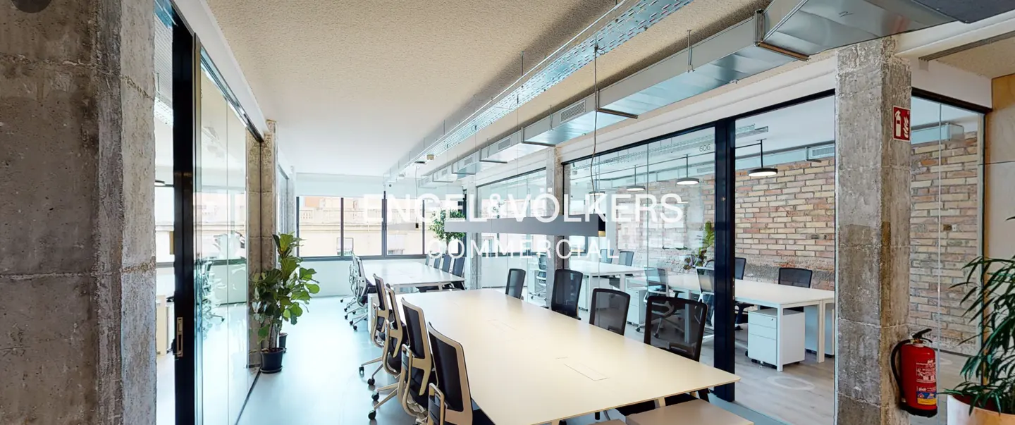 Bright office space with a long white table, black chairs, and glass-walled rooms. Concrete pillars and exposed ductwork add an industrial touch.