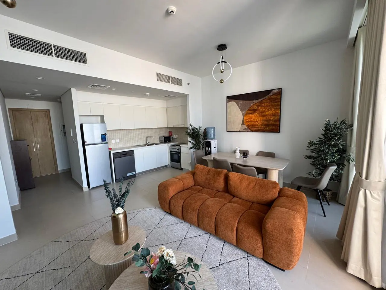 Bright, modern apartment interior with an orange sofa, white kitchen, and dining area. A patterned rug sits under round coffee tables.